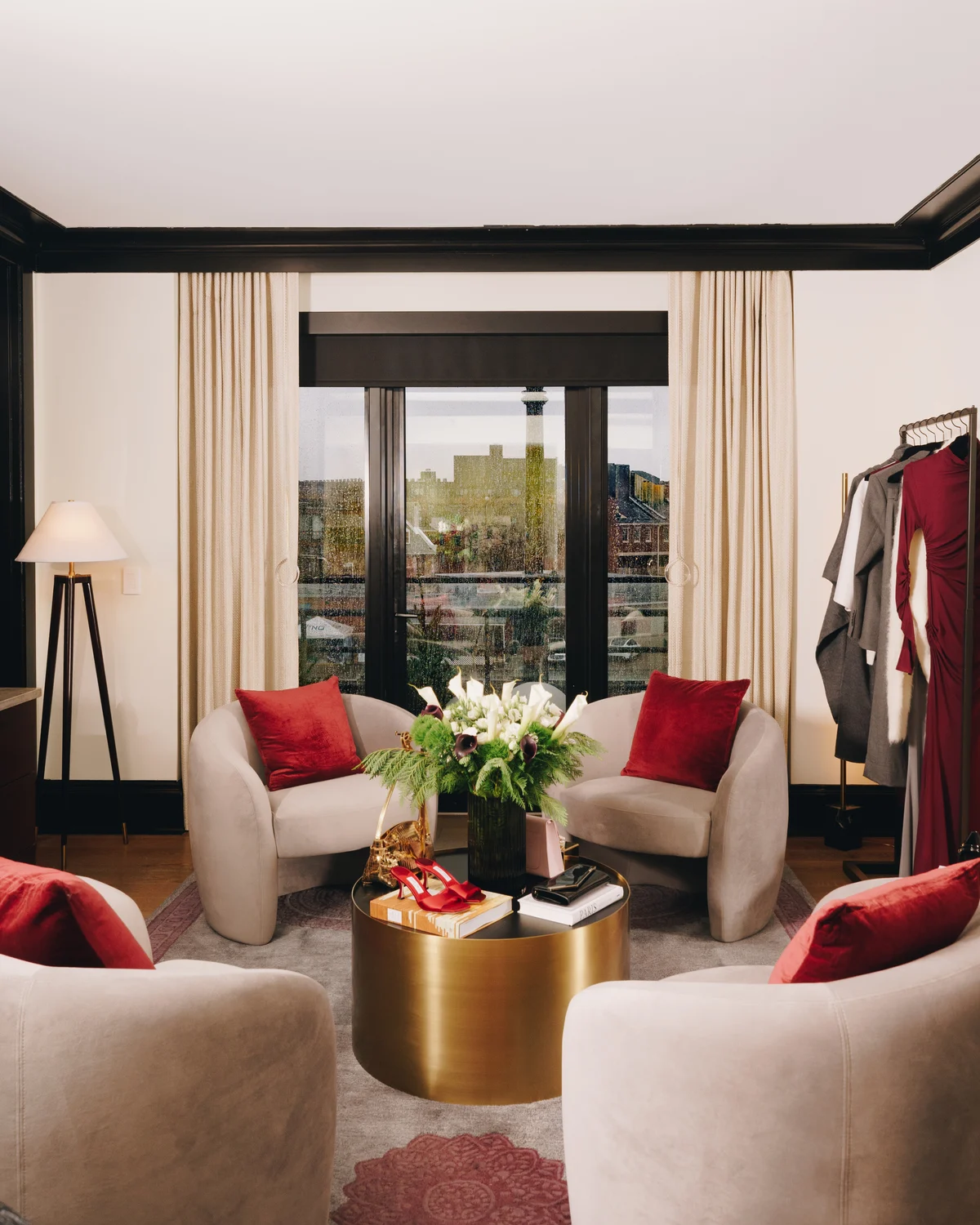 A suite sitting room with two cream armchairs and red cushions drawn around a brass drum table of flowers and gift boxes, a rail of clothing at the edge of the frame