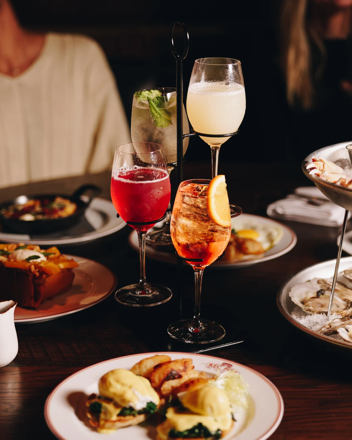 Four cocktails held in a black tiered stand, one clear with mint, one milky, one deep red and one orange over ice, with brunch plates and a bowl of oysters around them