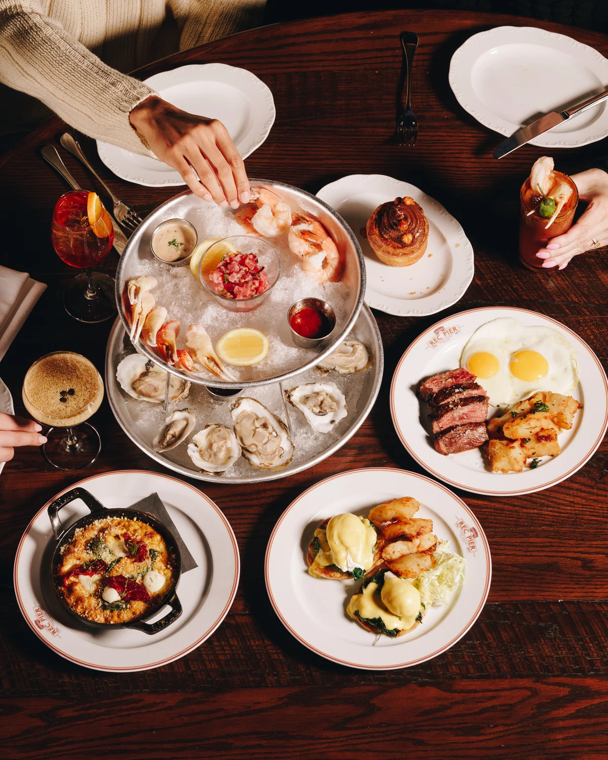 A table shot from above with a two tier seafood stand of oysters, shrimp and tuna tartare on ice, a steak and eggs plate, eggs benedict, a skillet of baked eggs and a hand reaching in for a shrimp