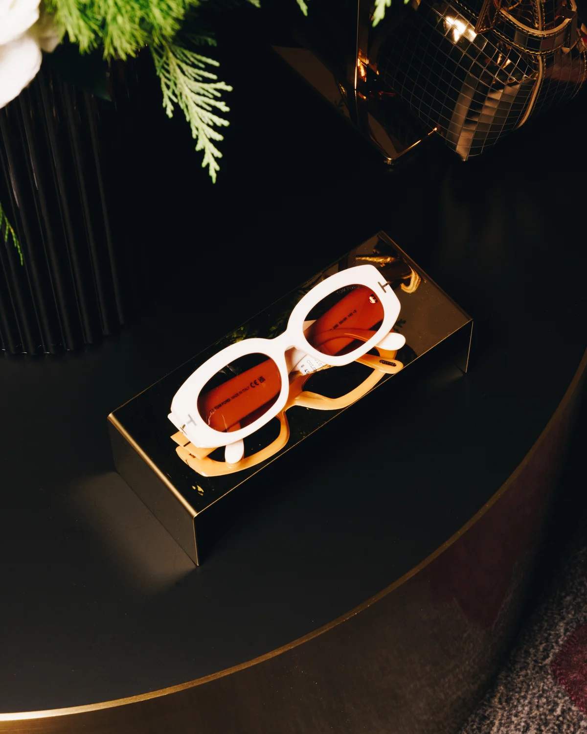 White sunglasses with orange-tinted lenses lying open in their gold case on a dark round table, greenery just above them