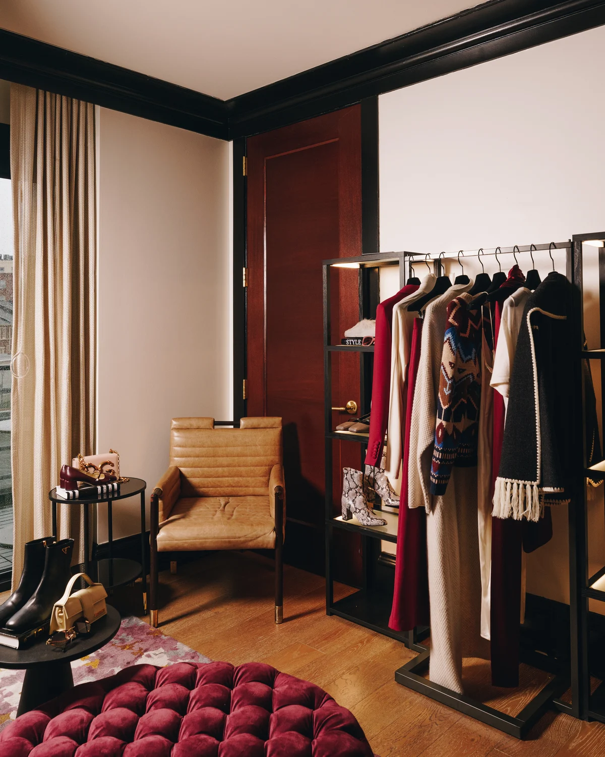 Rails of coats and knitwear standing in the middle of the room, a tan leather armchair and a deep red buttoned ottoman on the wood floor