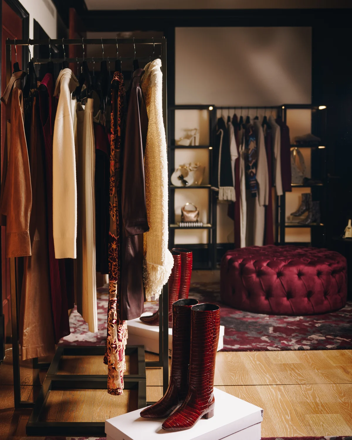 Tall burgundy crocodile boots on a low white plinth in front of rails of camel and cream coats