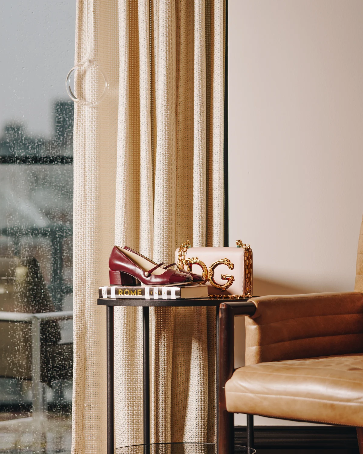 A burgundy heel and a blush leather bag with heavy gold hardware arranged on a book on a black side table, beside a rain-spotted window with the city behind it