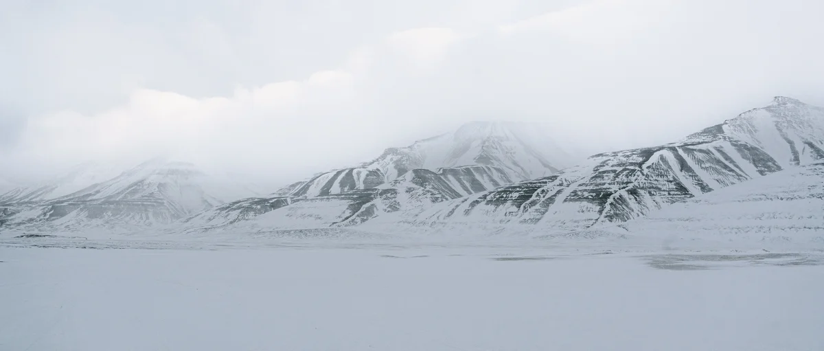 A long range of layered snow mountains under low cloud, seen across a flat frozen plain
