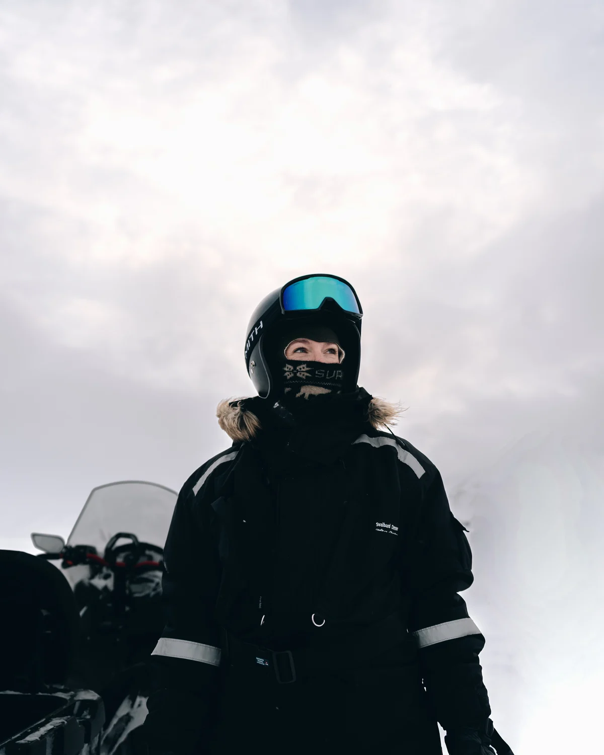 A guide in a helmet, mirrored goggles and a patterned face covering standing beside her snowmobile, looking off to one side