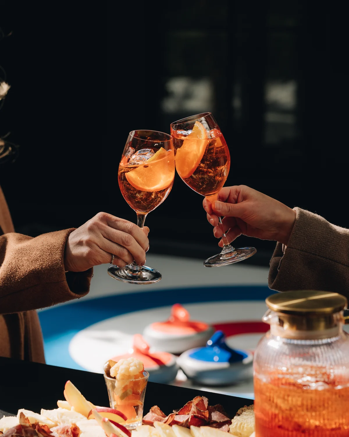 Two orange spritz glasses meeting over a spread of charcuterie and a glass drinks dispenser, the painted rings of the curling target and two stones just beyond