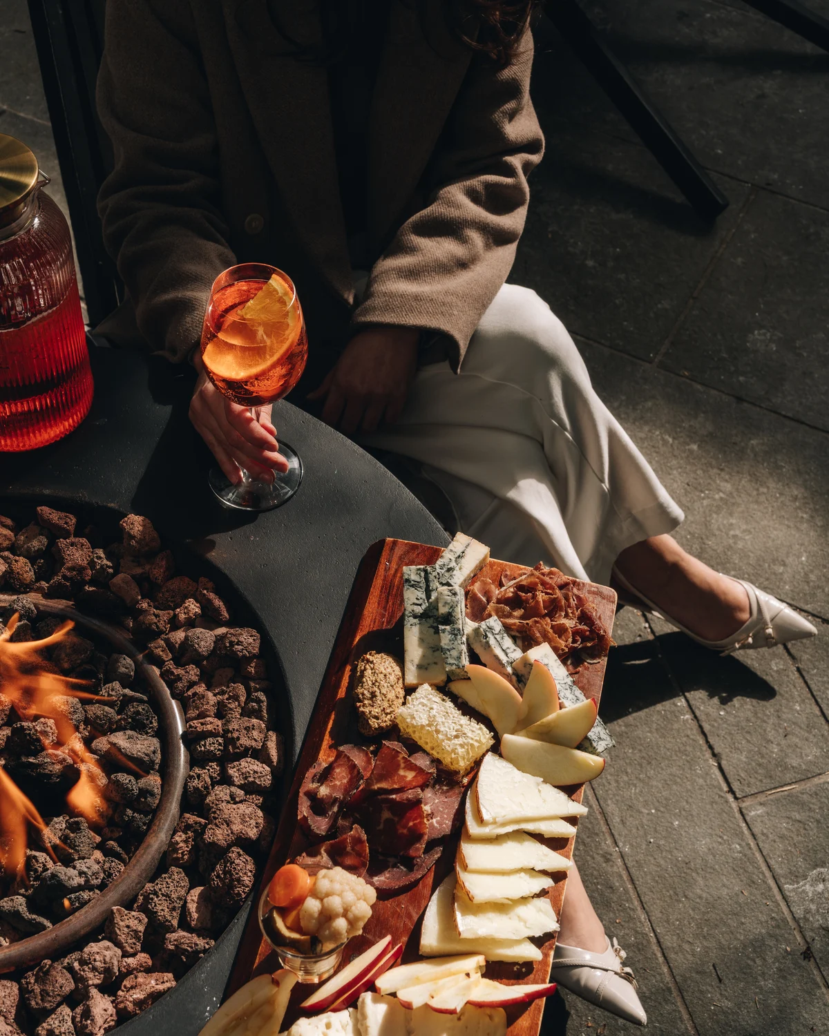 Looking down on a guest in a camel coat holding a spritz, a long board of cheese, cured meat and fruit beside a lit fire pit