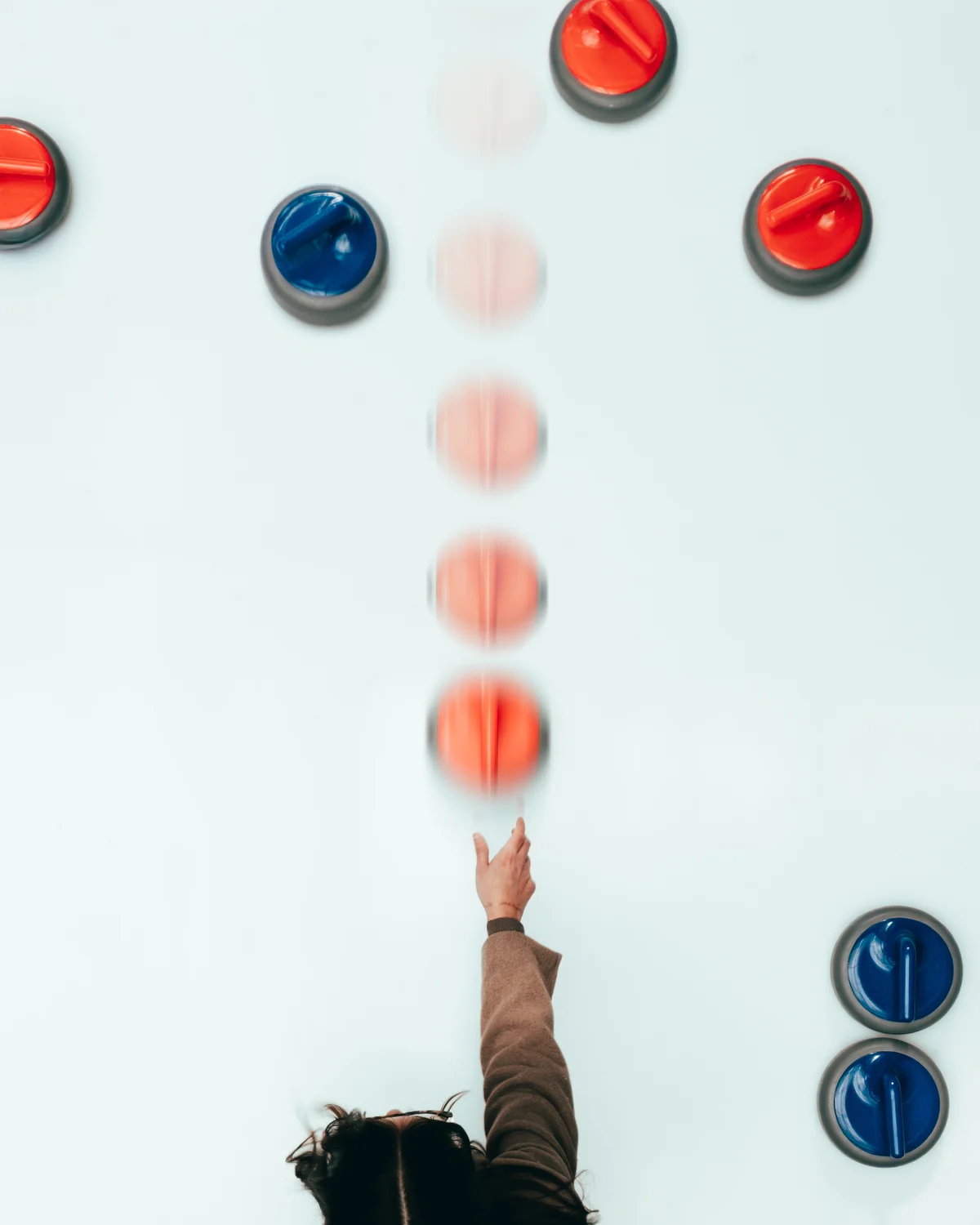 From directly overhead, a curler's arm still extended after the release and the red stone repeating up the ice in a long exposure, other stones at rest around the edges