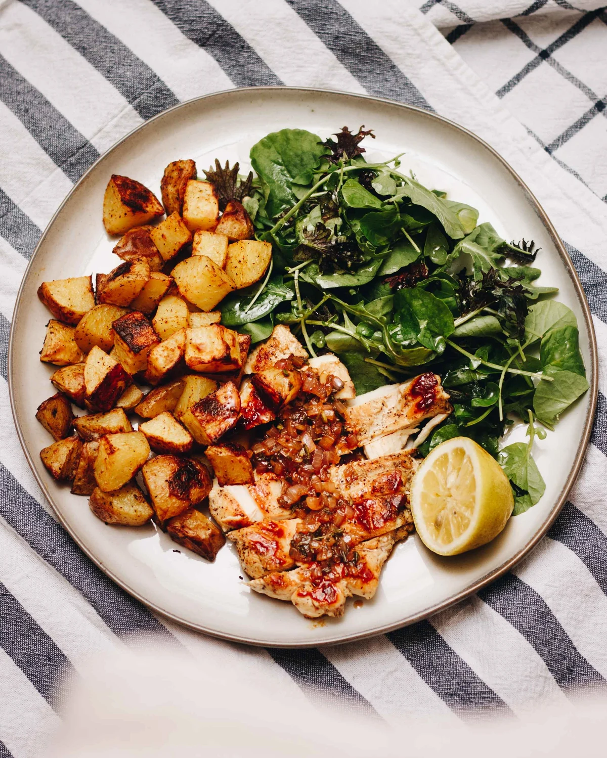 Sliced chicken with chilli and shallot dressing, roast potatoes, dressed leaves and half a lemon on a stoneware plate, on a striped cloth