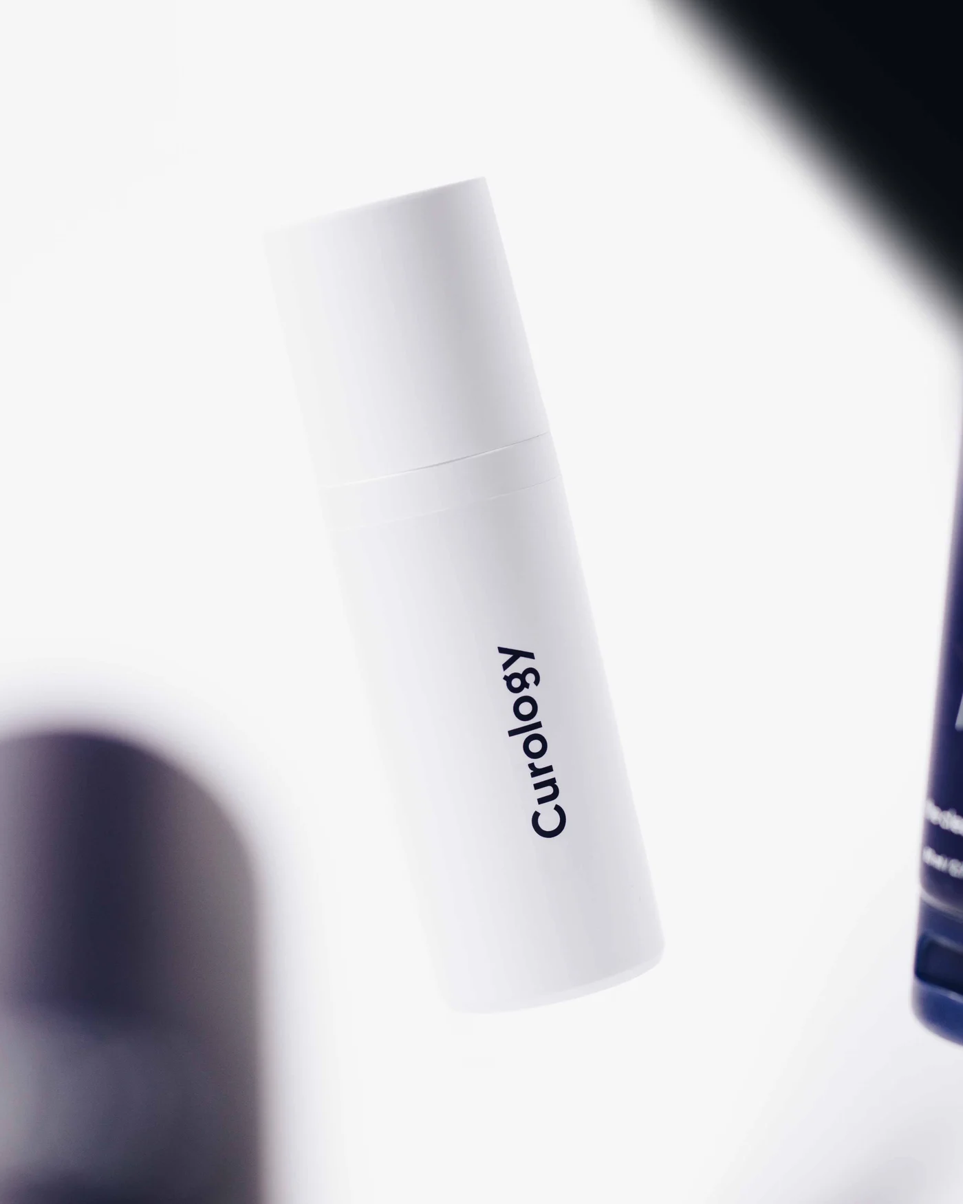 A white cylindrical skincare bottle tipped at an angle on white, soft out-of-focus shapes crossing the front of the frame