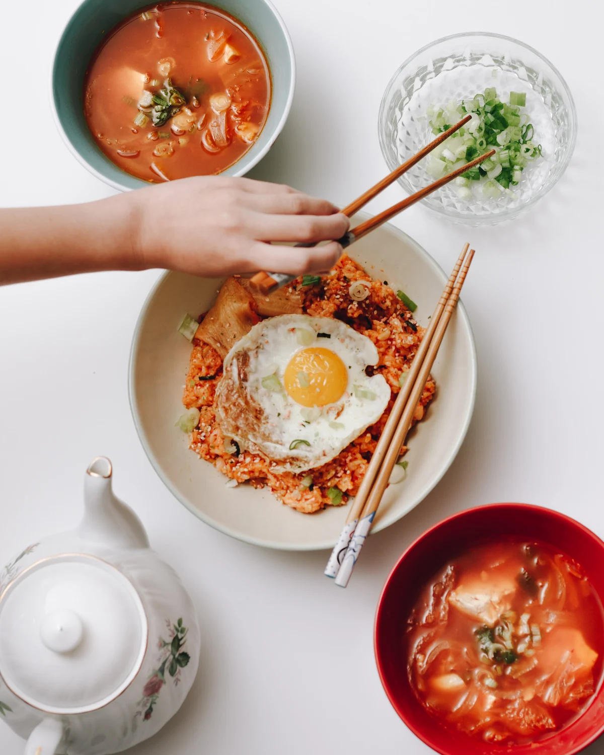 A bowl of kimchi fried rice under a fried egg, a hand reaching in with chopsticks, with two bowls of stew, a dish of spring onion and a floral teapot around it