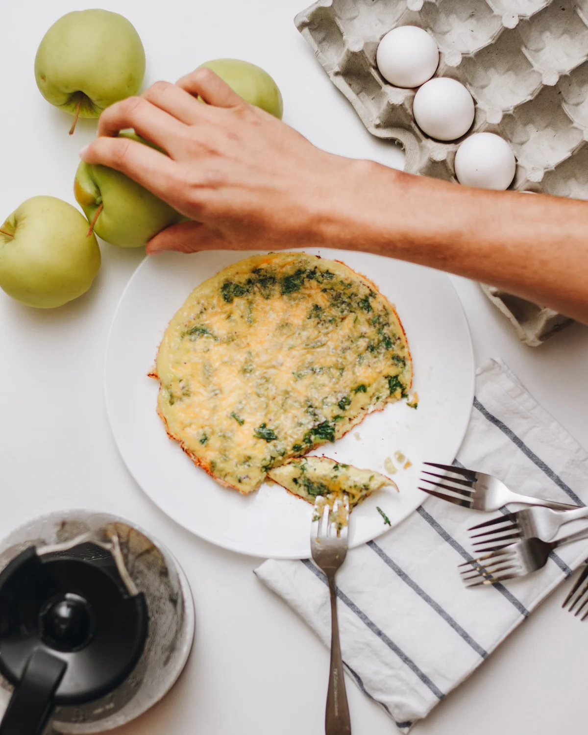 A spinach and cheese frittata on a white plate with one slice cut, a hand steadying green apples beside it and a carton of eggs above