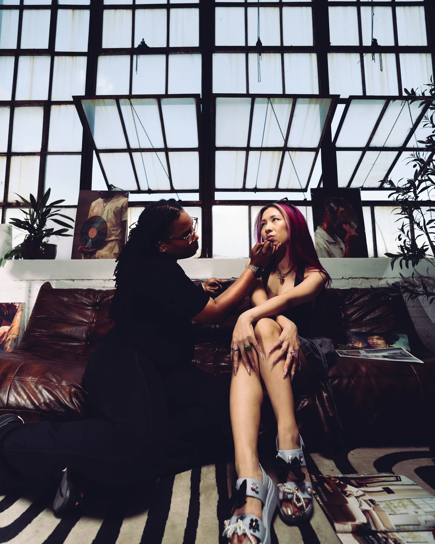 Two women on a leather sofa under a wall of loft windows, one leaning in to talk