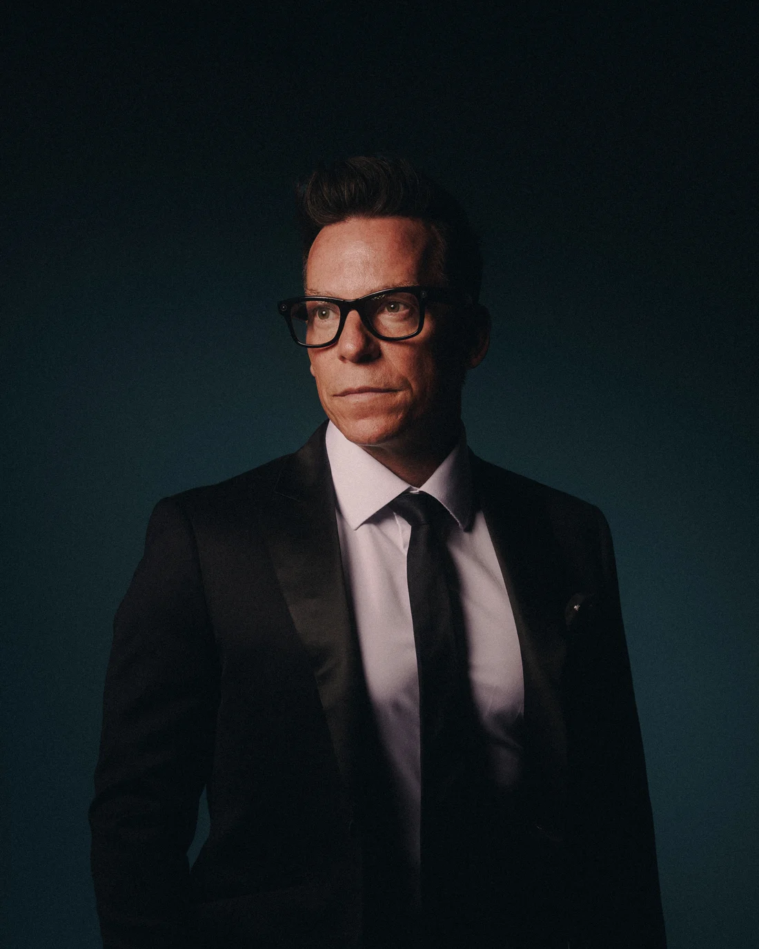 A man in a tuxedo and heavy black glasses against deep teal, looking straight down the lens