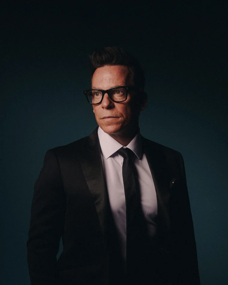 A man in a tuxedo and heavy black glasses against deep teal, looking straight down the lens
