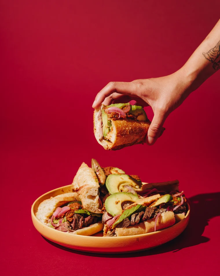 A tattooed hand lifting a loaded sandwich off a coral plate stacked with two more, on scarlet