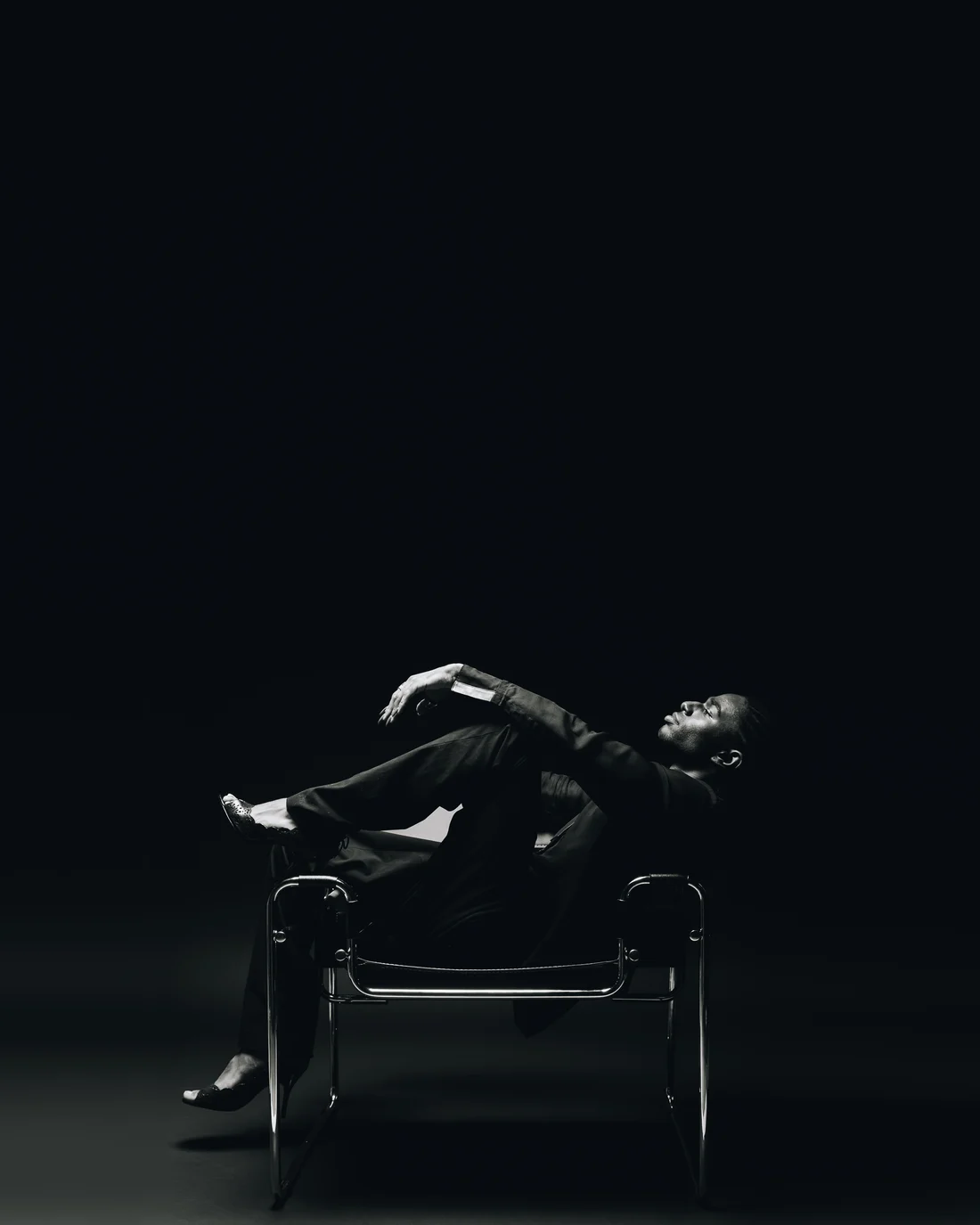 A man in a black suit thrown back across a tubular steel sling chair in a small pool of light