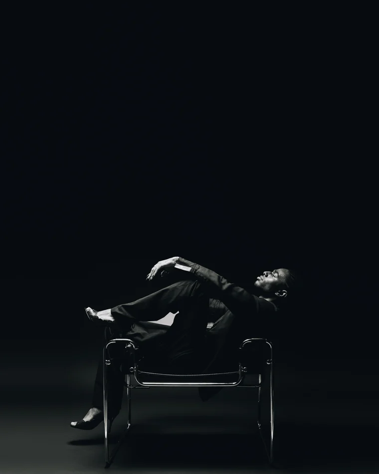A man in a black suit thrown back across a tubular steel sling chair in a small pool of light