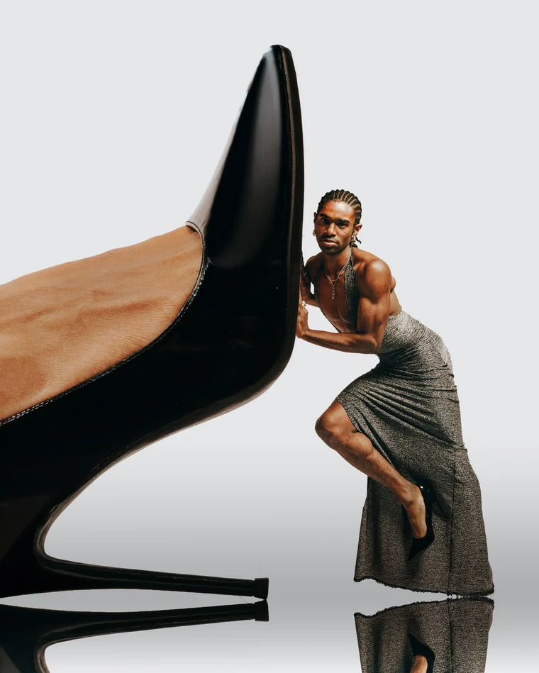 A dancer in a grey draped dress leaning their whole weight into an enormous black stiletto, as if trying to push it over