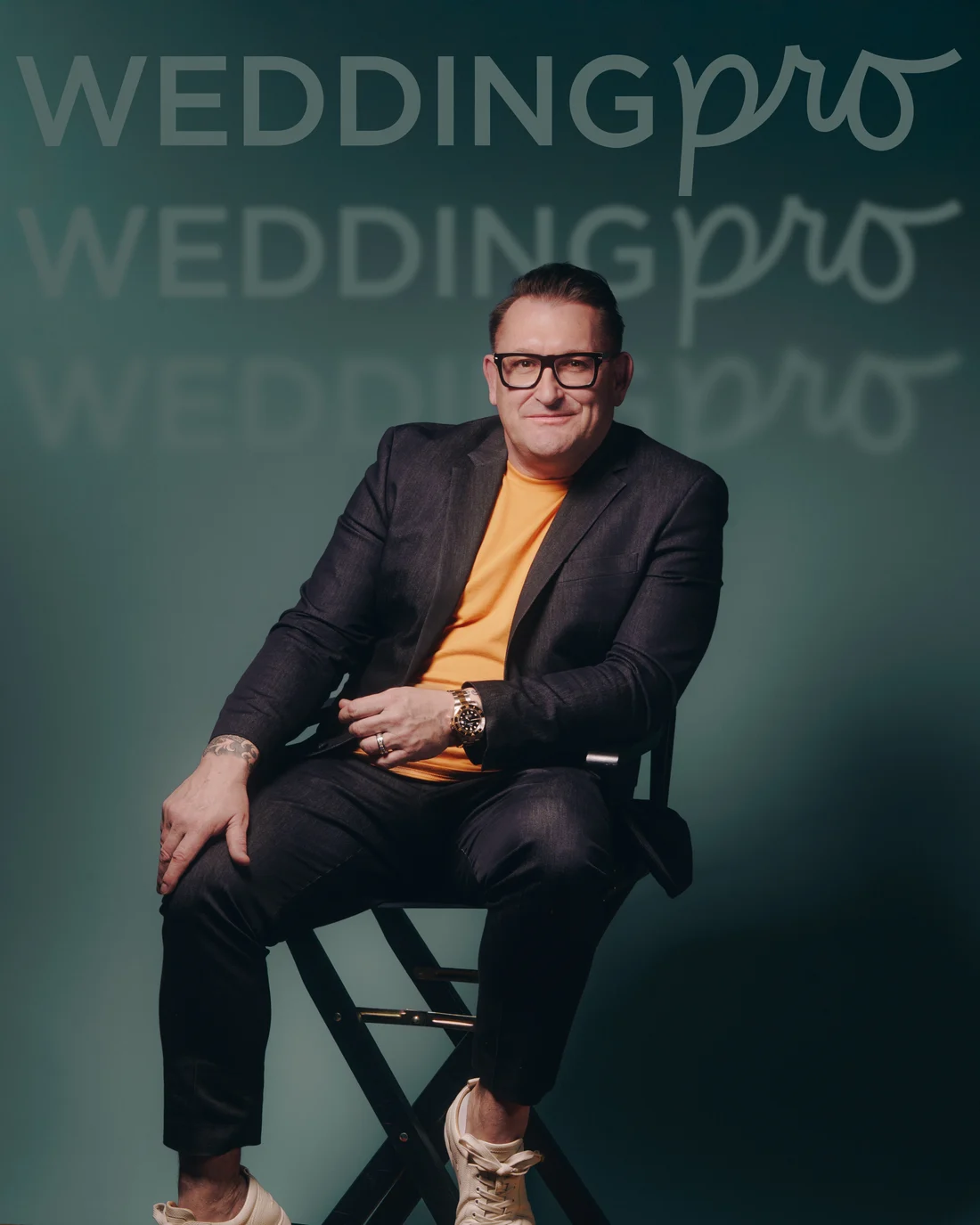 The same man seated on a director's chair with his legs crossed, against a teal wall of repeated brand lettering