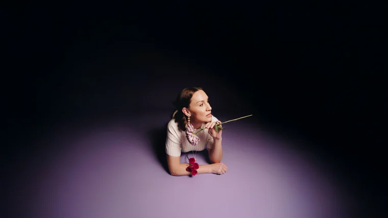 A woman lying on her front in a pool of lilac light, chin resting on her hands, a red bloom pinned at her collar