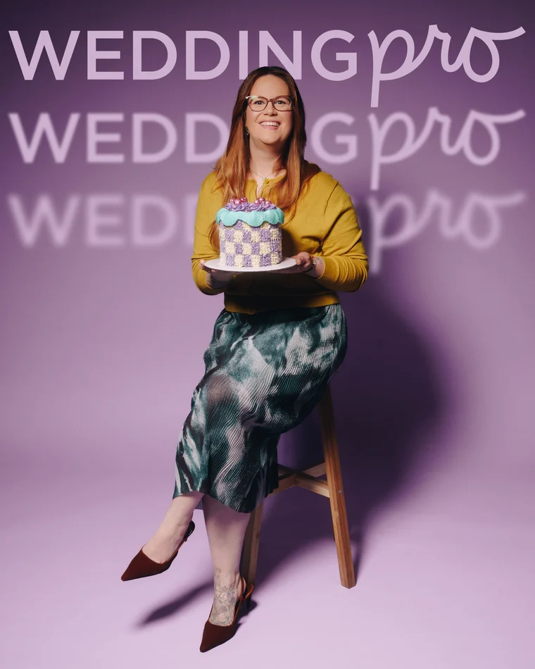 A woman in a mustard cardigan and painted silk skirt perched on a stool, holding out a chequerboard cake on a tray