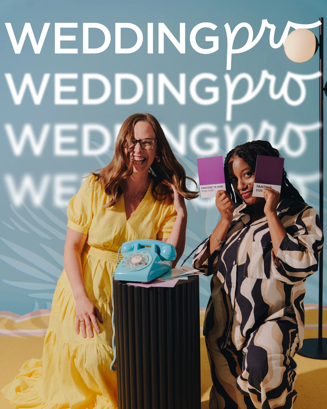 Two women either side of a fluted plinth with a teal rotary telephone on it, one leaning in a yellow dress, the other holding up purple swatch cards