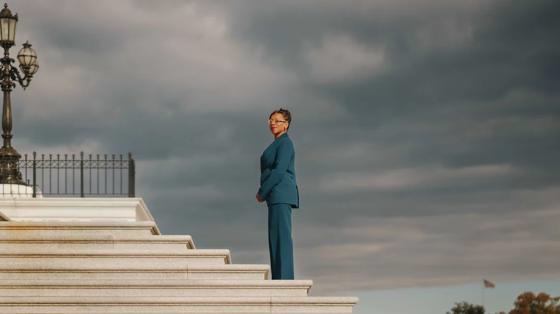 A woman in a teal suit standing alone at the top of a wide flight of marble steps under heavy grey cloud, an ornate cast iron lamppost at the edge of the frame
