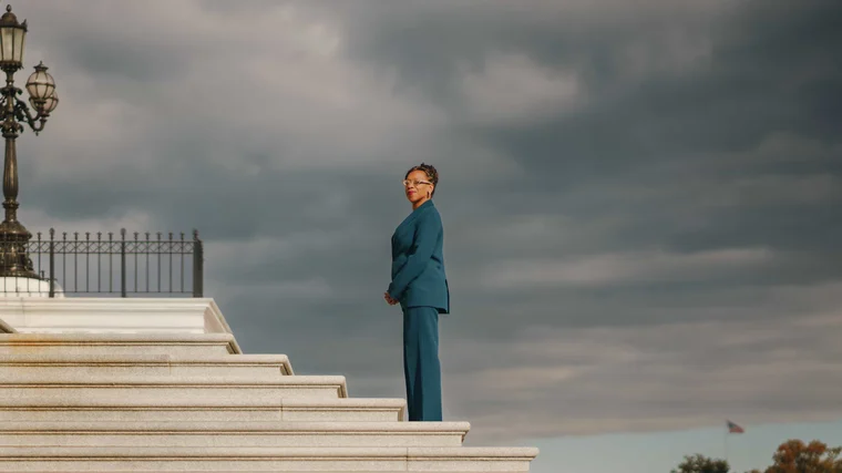 A woman in a teal suit standing alone at the top of a wide flight of marble steps under heavy grey cloud, an ornate cast iron lamppost at the edge of the frame