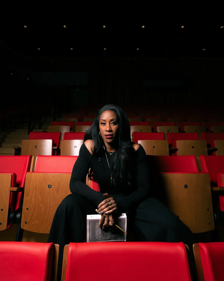 A woman in black sitting alone in a row of red theatre seats in an empty auditorium, a book in her lap
