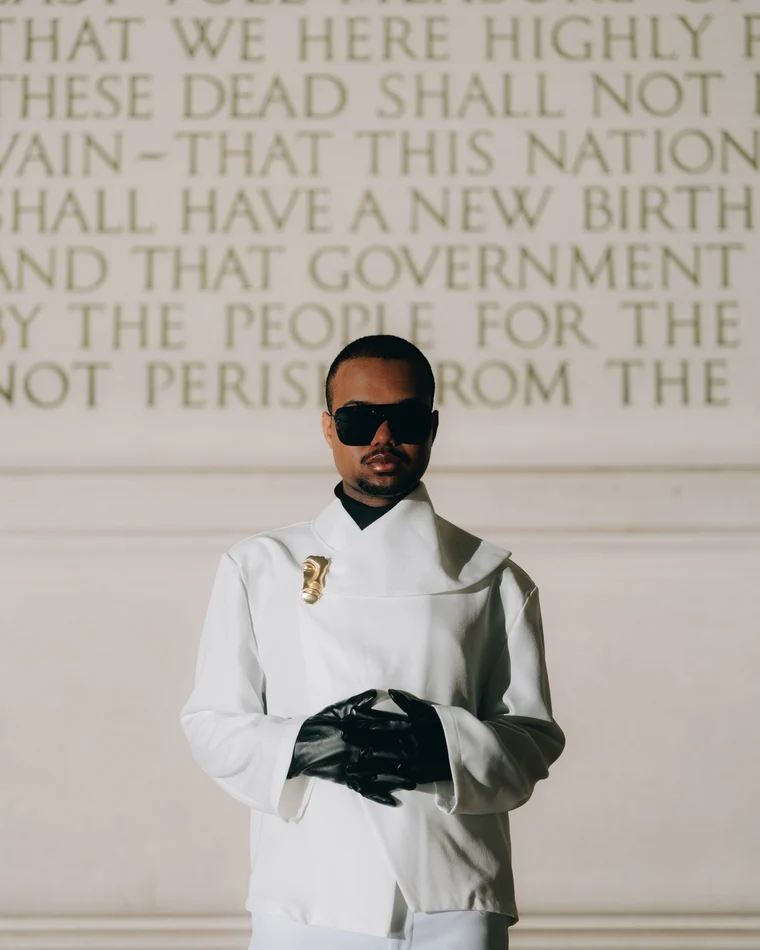 A figure in a sculptural white jacket and one long black glove, standing against a stone wall carved with the closing lines of the Gettysburg Address