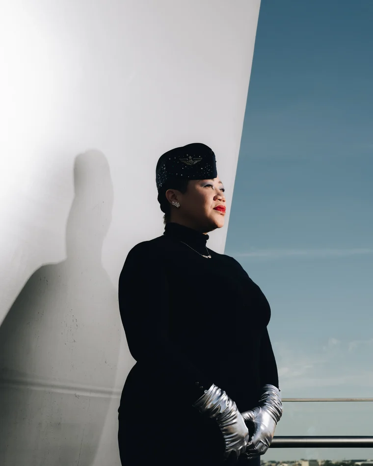 The same woman in black with silver opera gloves, in profile against a white curved wall that throws her shadow twice her size