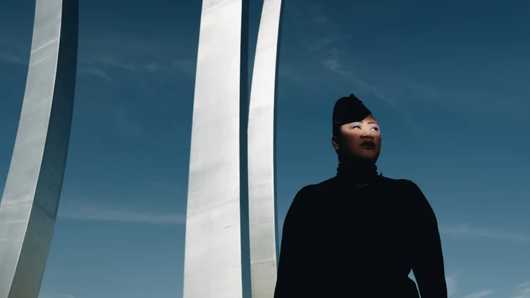 A near silhouette in a black turtleneck and beaded cap looking up, framed by three soaring white spires against open blue sky