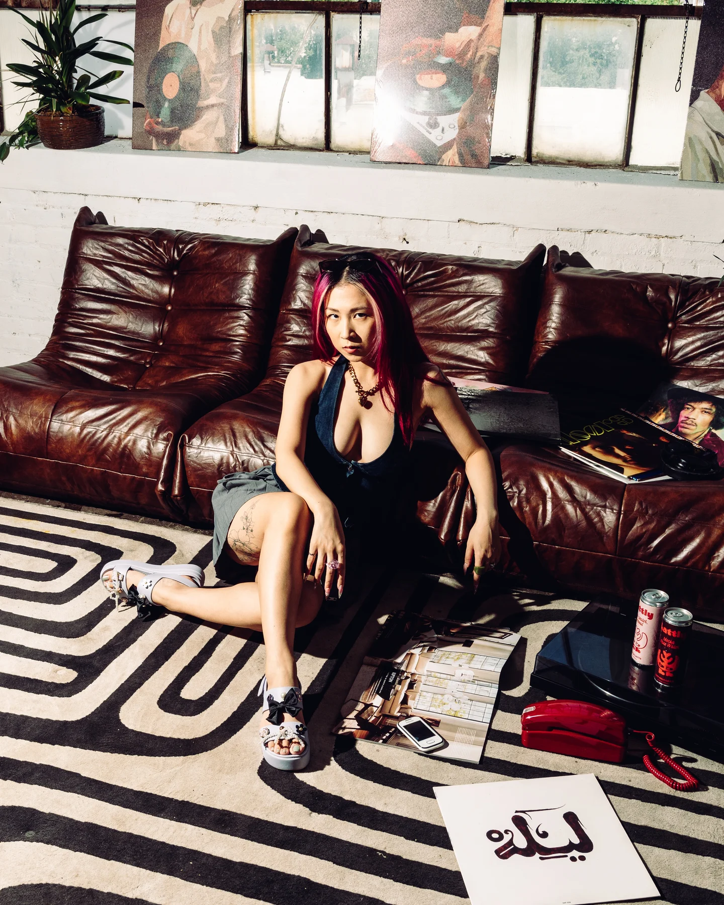 The woman with magenta hair on the leather sofa in a black halter, cans at her feet and a card on the rug