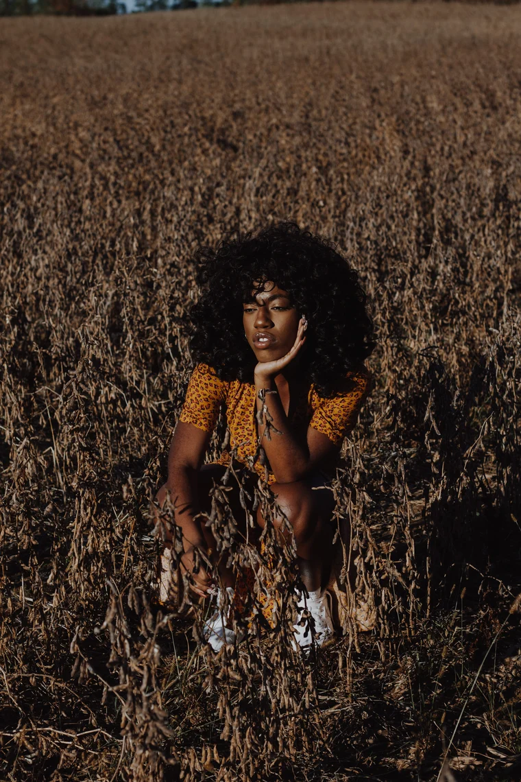 A woman with an afro crouched low in a harvested field, chin resting on her hand