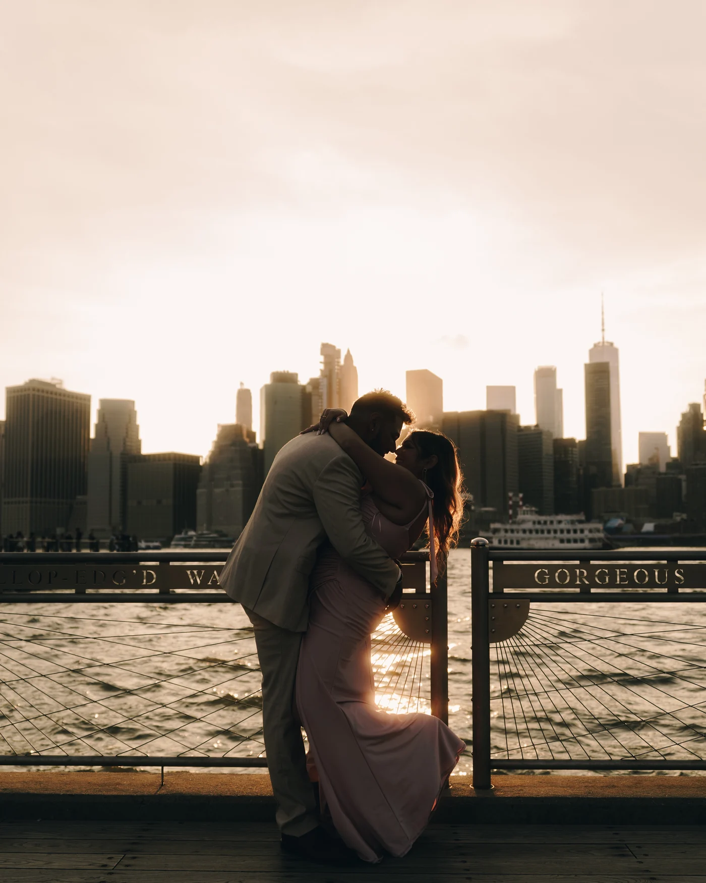 A couple kissing against the railing of a waterfront promenade, the towers of lower Manhattan going gold behind them