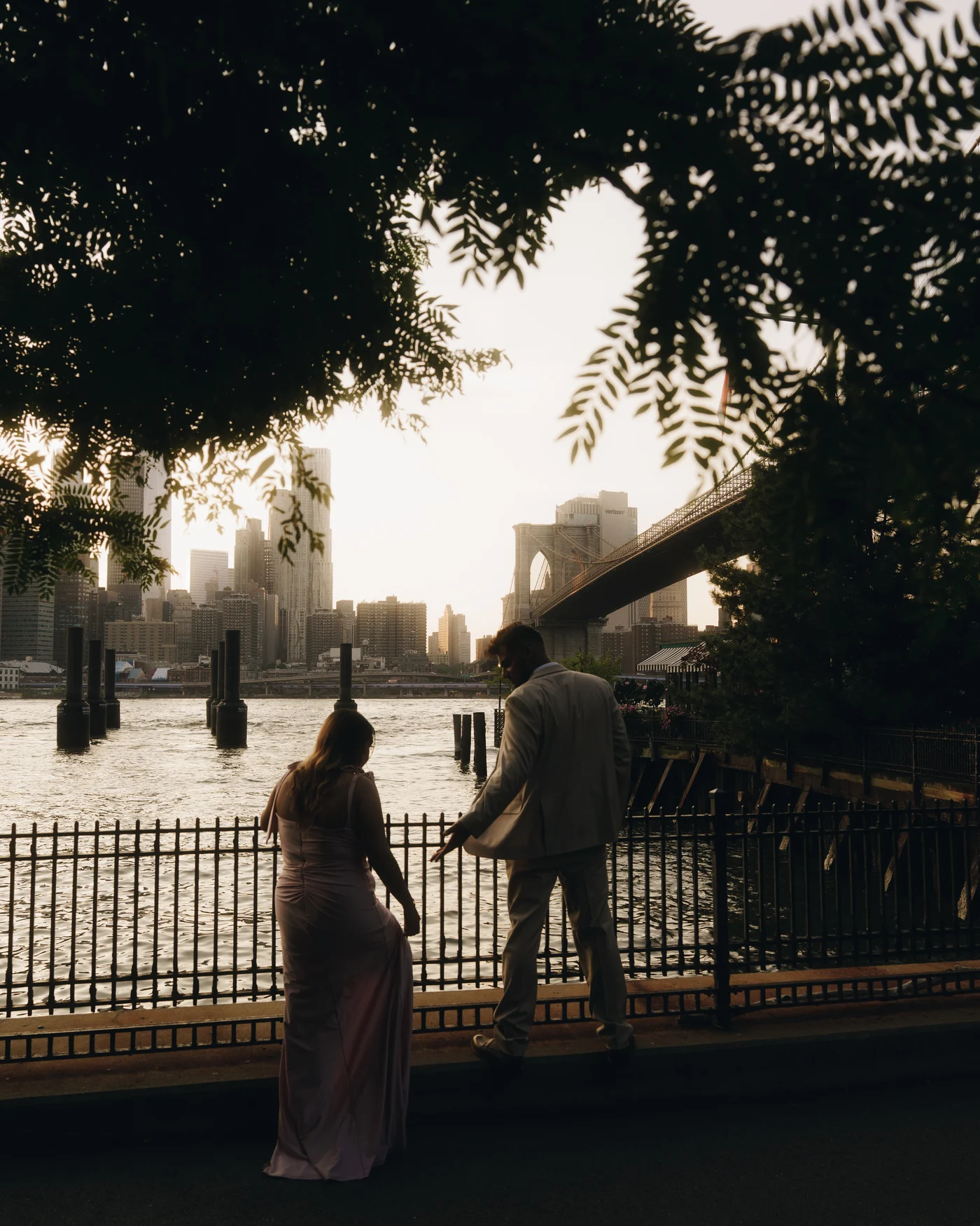 Seen through overhanging leaves, a couple at an iron railing above the water with the bridge and the skyline beyond