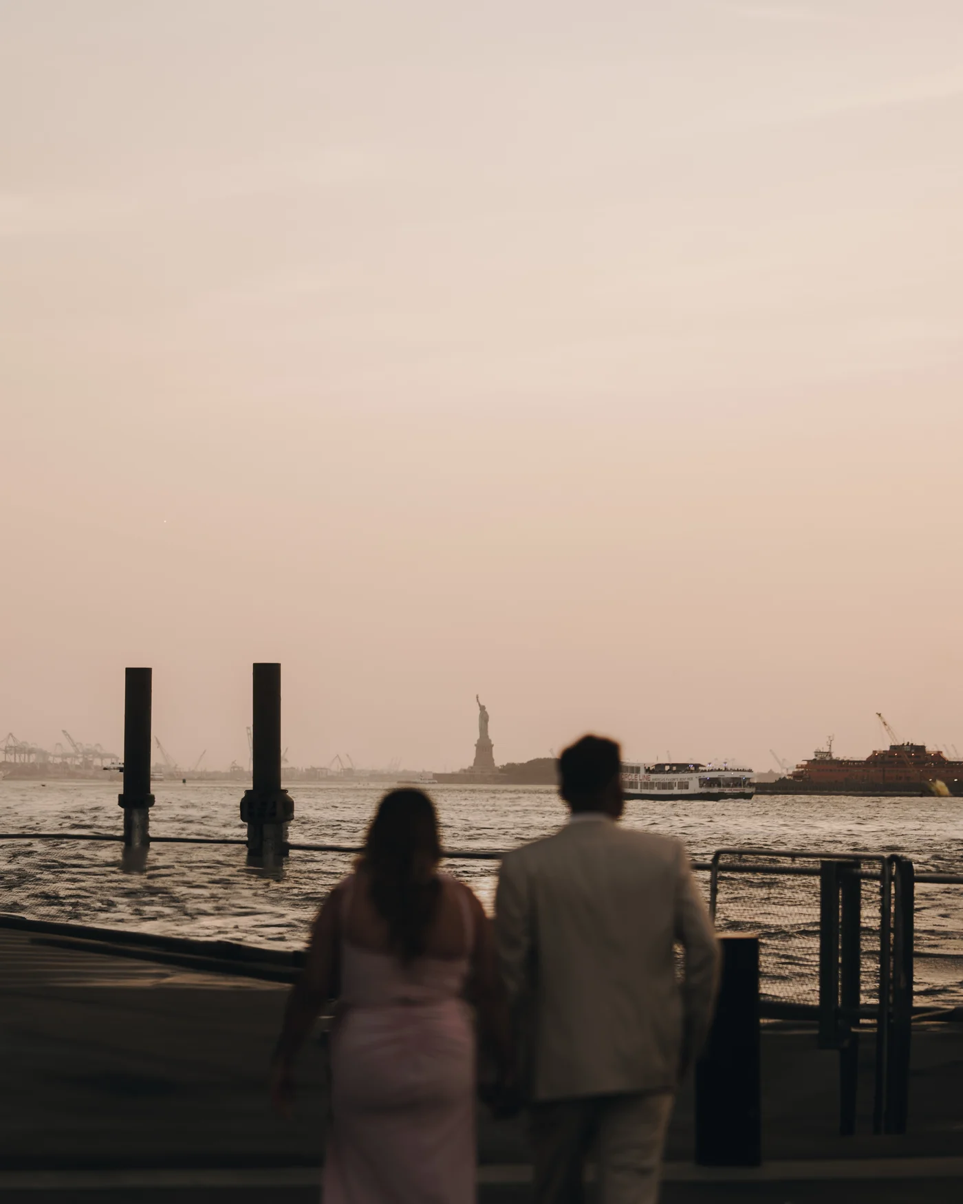 A couple from behind at the water's edge, holding hands and looking out toward the Statue of Liberty