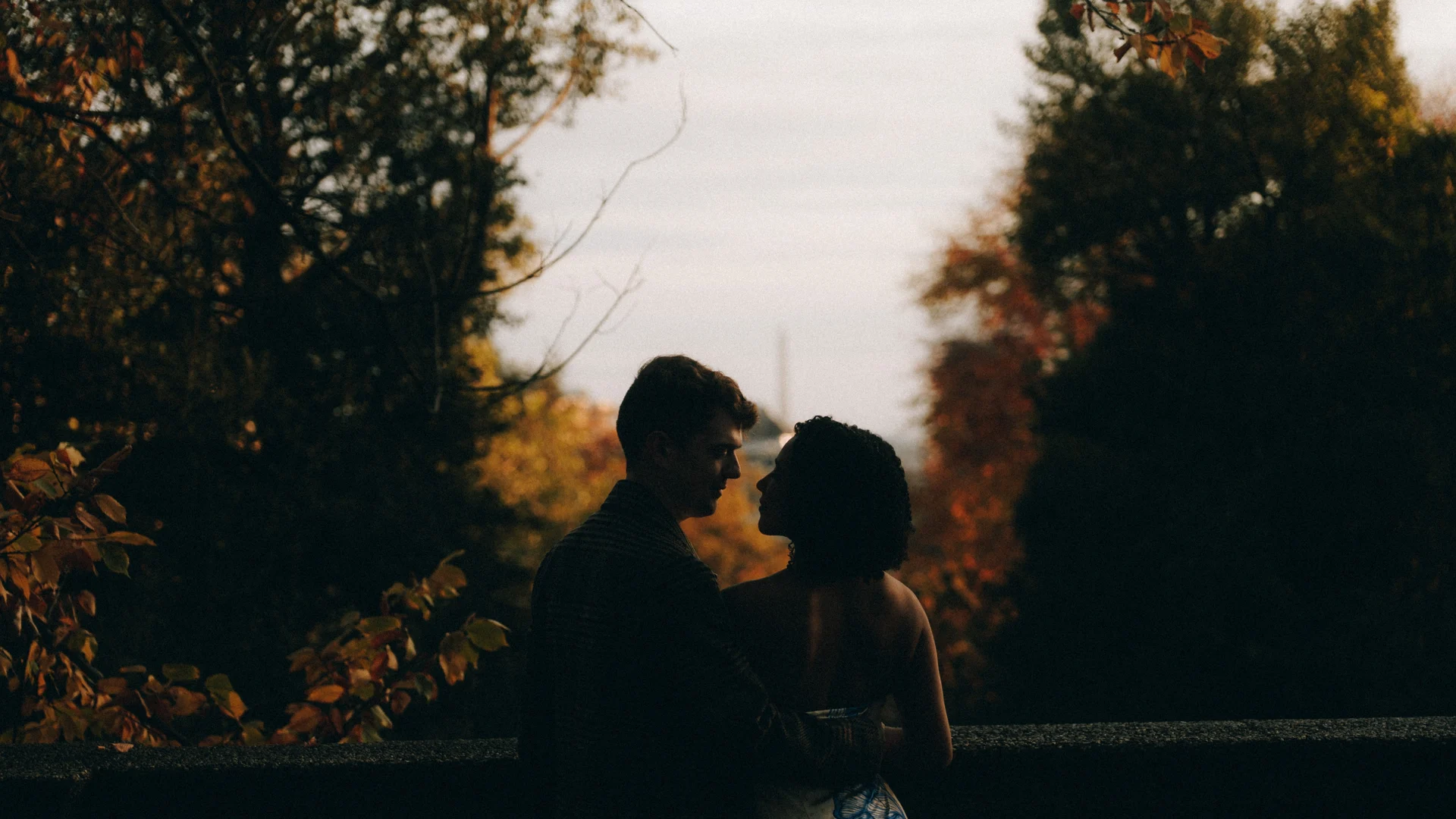 A couple in silhouette facing each other at a stone parapet at dusk, autumn trees on both sides and the Washington Monument a pale needle on the horizon