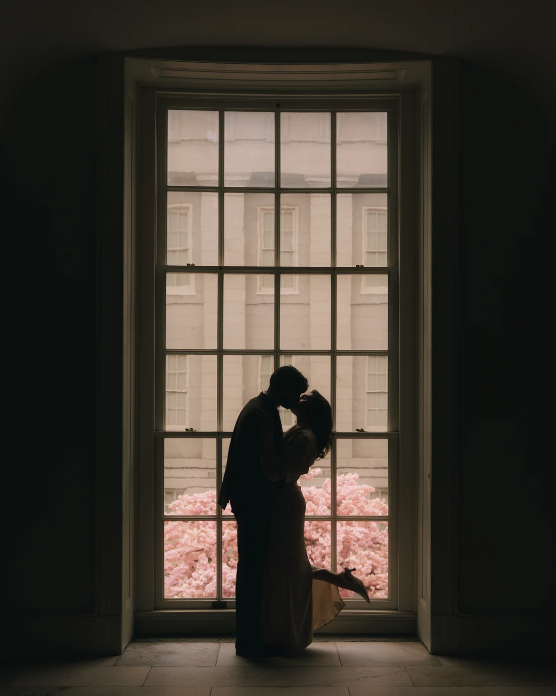 A couple silhouetted in a tall arched museum window with cherry blossom glowing pink outside it