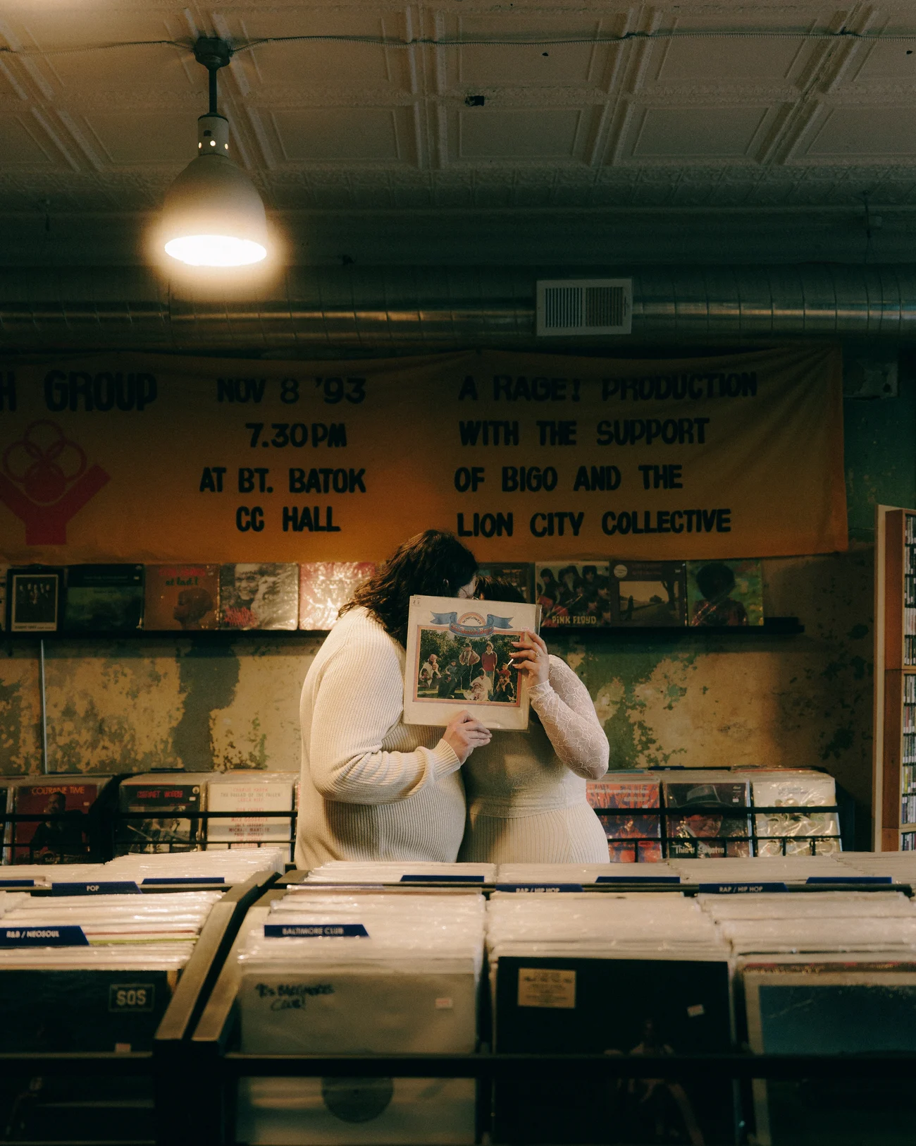 In a record shop, one of them holds an album sleeve up over their face while the other leans in behind the crates