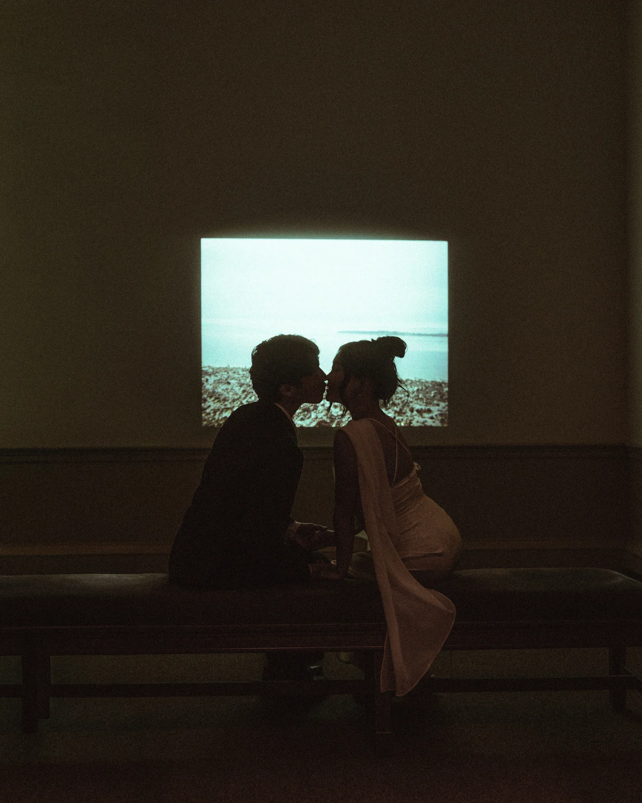 A couple close together on a bench in a dark room, lit only by the glow of a video projection on the wall