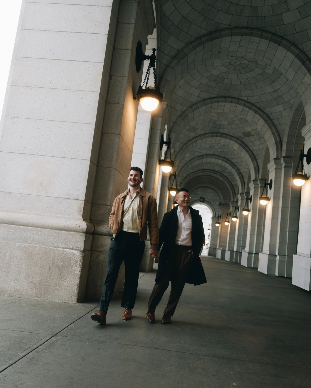 A couple walking hand in hand through a long vaulted stone arcade under hanging lamps