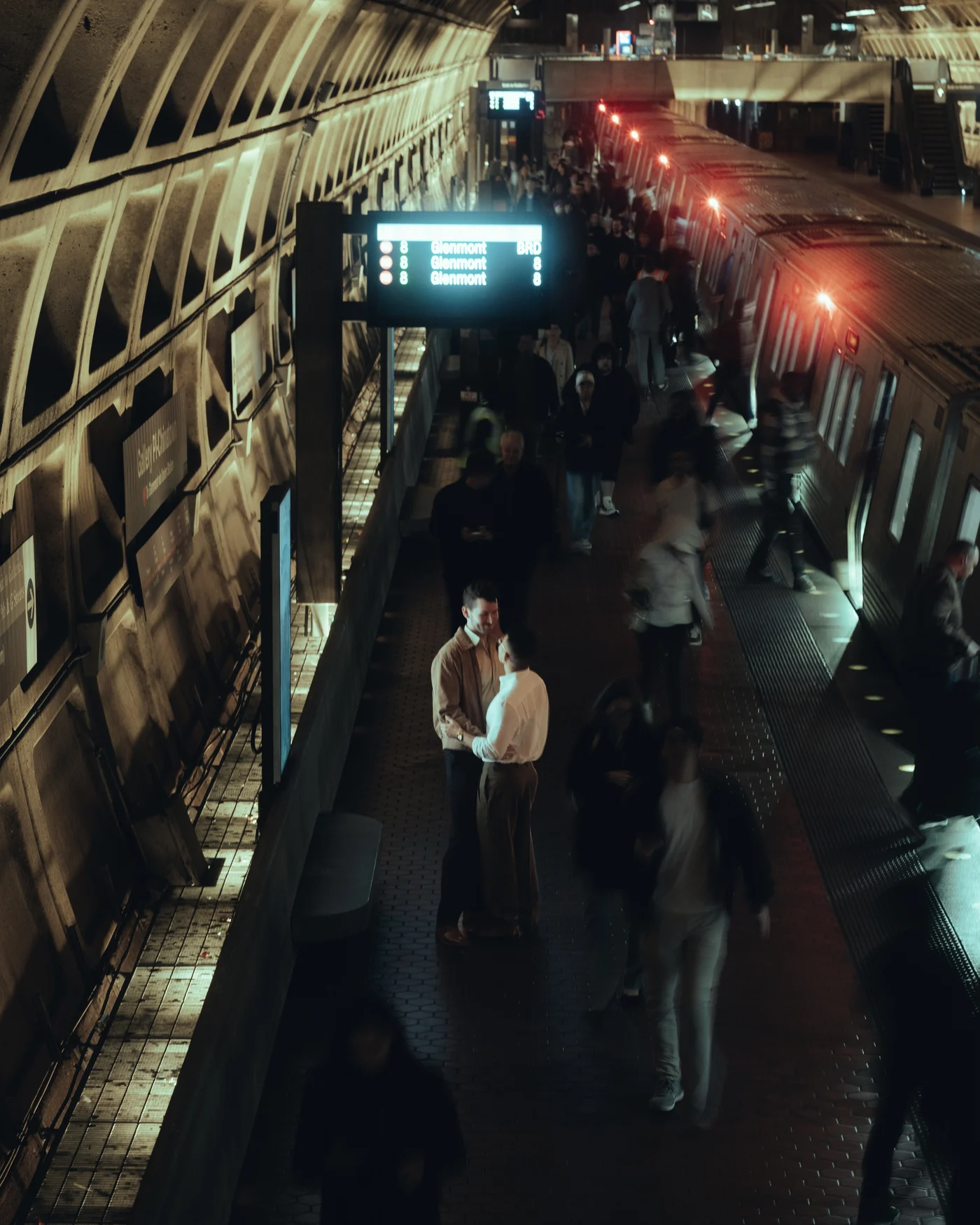 From above, a couple standing still on a Metro platform while the evening crowd blurs past them