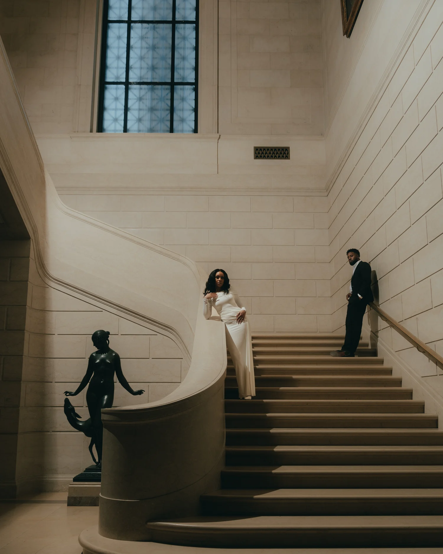 A pale marble stairwell with a bronze figure on its plinth, a woman in white on the landing and her partner on the steps above