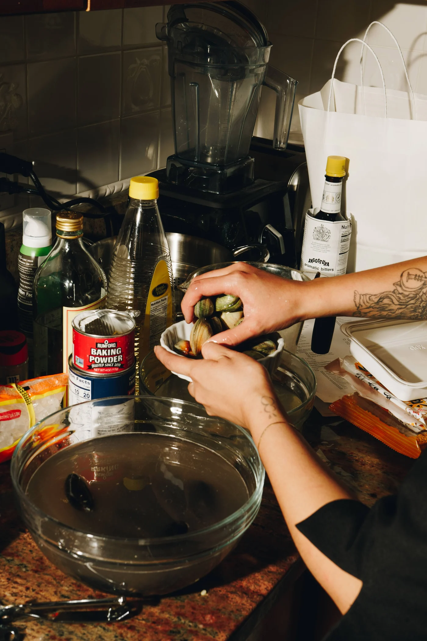 Tattooed hands sorting clams over a bowl in a crowded home kitchen