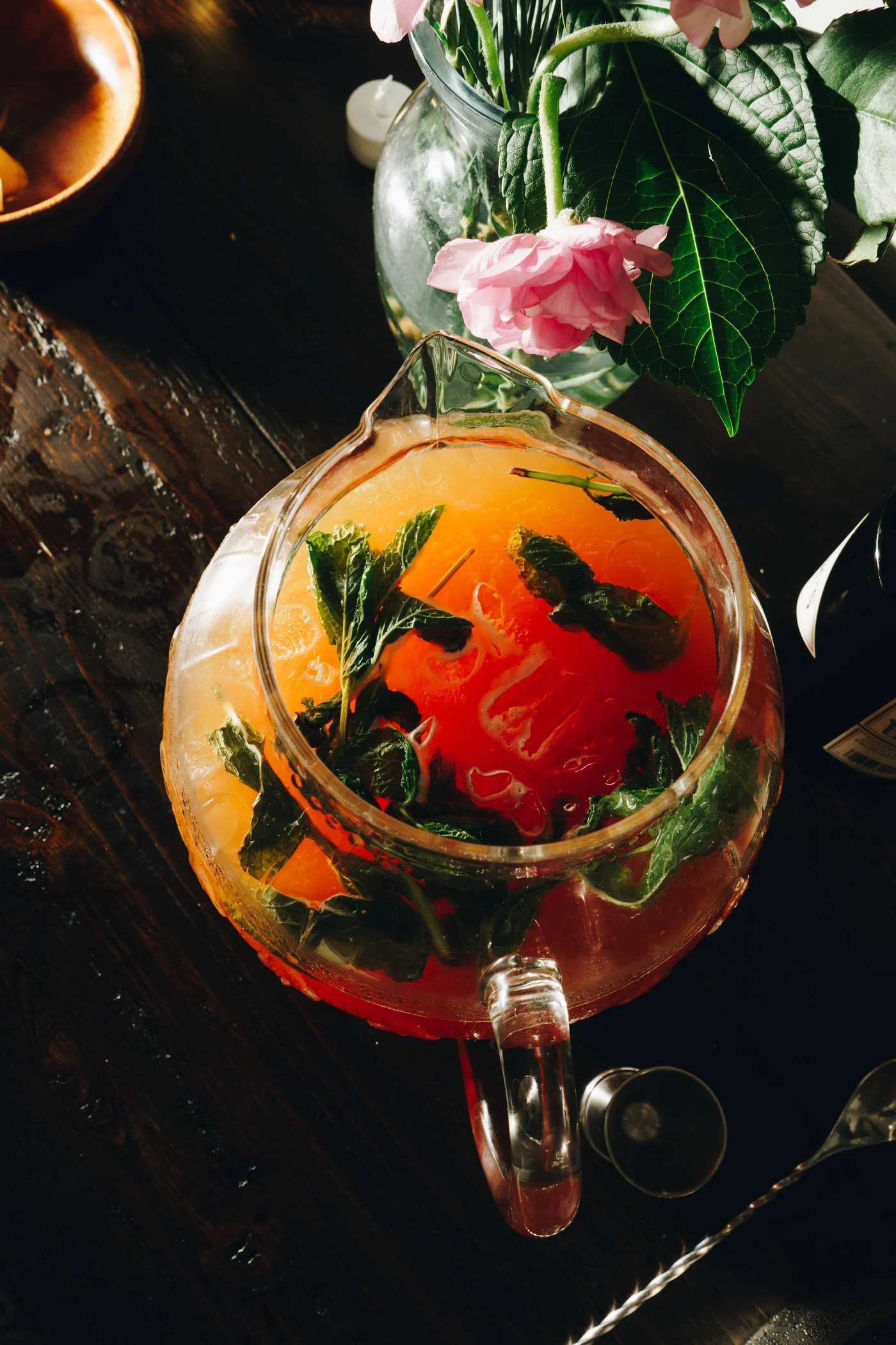 A glass pitcher of coral punch thick with mint leaves, pink roses leaning overhead