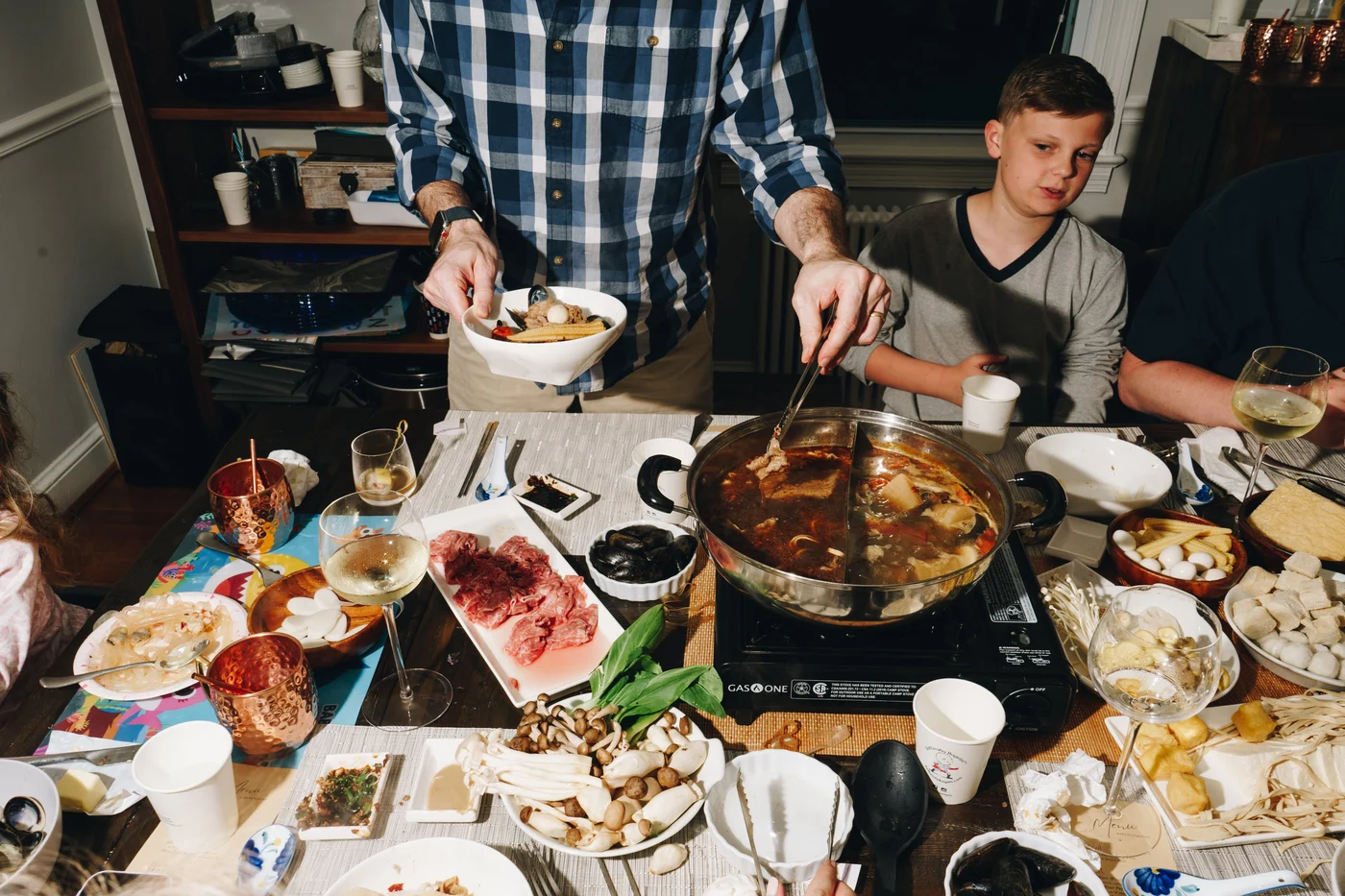 A divided hot pot simmering mid table while a boy watches his bowl being filled