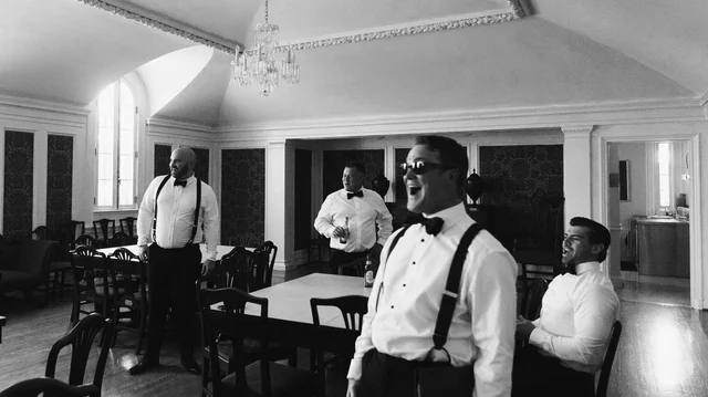 Groomsmen in suspenders and bow ties share a laugh in a vaulted getting-ready room beneath a crystal chandelier