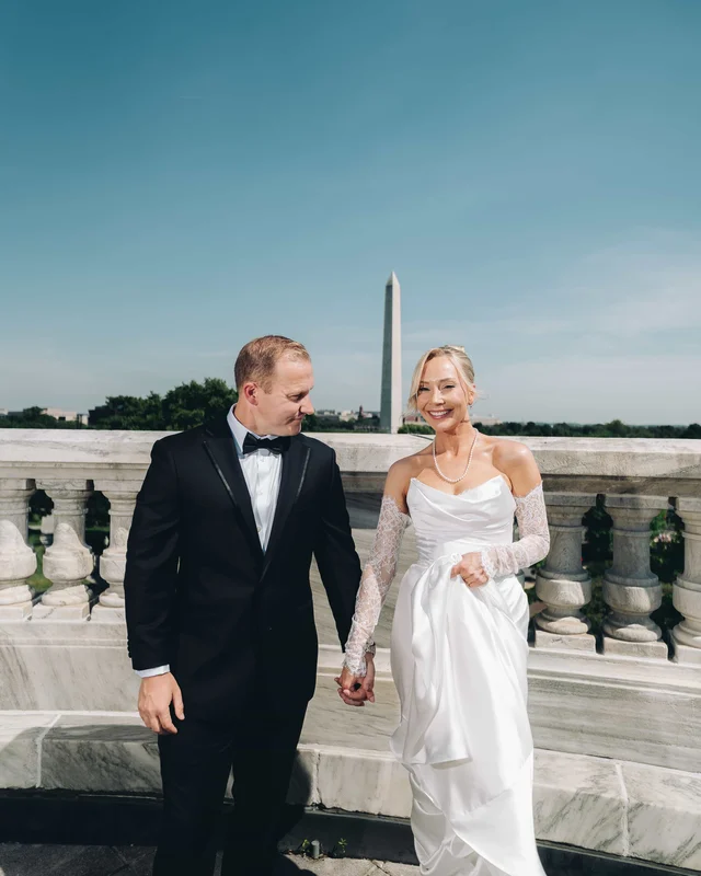 The bride and groom hold hands on a marble rooftop balustrade, the Washington Monument rising behind them under a clear sky